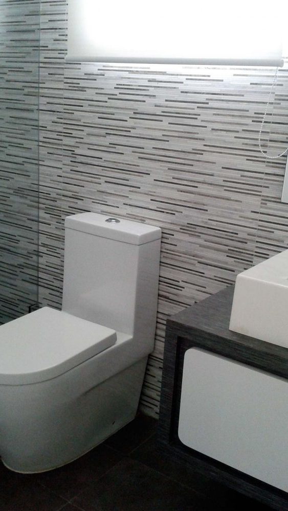baño con pared de porcelanato de rayas horizontales de distintos tonos de grises, lavamanos rectangular con mueble laminado color wengue
